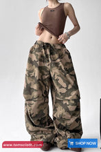 Person wearing camouflage pants and a brown top on a white background with 'TomsCloth' branding.
