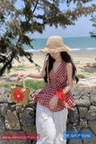 Woman on a beach wearing a red checkered top and white pants, holding a straw bag with a flower design.