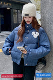 Woman wearing a blue puffer jacket and white beanie leaning against a wall.