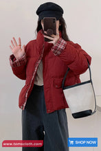 Person wearing a red puffer jacket and black cap, holding a phone, with a visible brand logo at the bottom.