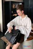 Woman in a white blouse opening a black quilted handbag in a room with shelves in the background.