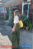 Woman in green pants and white shirt with yellow bag walking on a street, with a shop and plants in the background.