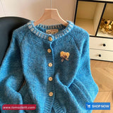 Blue knitted cardigan with a heart-shaped brooch on a hanger, brand 'Toms Cloth' visible.