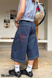 Person wearing blue checkered shirt and denim shorts with a beige bag, standing indoors.