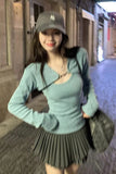 Woman wearing a light blue sweater and pleated skirt with a dark cap in an urban setting.