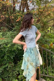 Person wearing a light blue top and floral skirt standing outdoors with greenery in the background