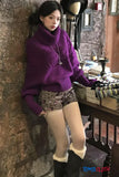 Person wearing a purple sweater and patterned shorts leaning against a stone wall.