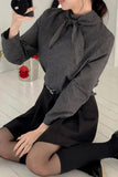 Person wearing a checkered blouse with a large bow and black skirt sitting on the floor.