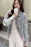 Woman wearing a light gray floral embroidered jacket holding a white cup outdoors.