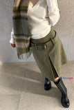 Person wearing a white sweater, green skirt, and black shoes against a concrete wall.