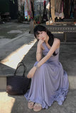 Woman in a purple dress sitting on the ground with a woven bag next to her, in an outdoor setting.