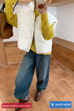 Person wearing a white puffer vest over a yellow sweater with blue jeans and brown boots, standing in a room with wooden flooring.