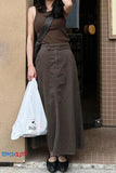 Woman wearing a brown sleeveless top and long skirt, holding a white plastic bag, standing against a tiled wall.