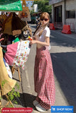 Woman in a white top and red checkered skirt standing on a street with a vendor's stall in the background.