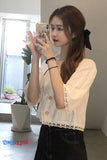 Woman taking a mirror selfie wearing a white lace top and black pants in a room with plants.