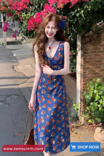 Woman in a blue floral dress standing outdoors with flowers and a brick wall in the background.