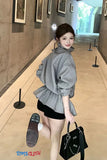 Woman in a gray sweater and black skirt holding a black handbag in an indoor setting.