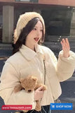 Woman in a cream-colored coat and hat holding a teddy bear, with a blurred background.