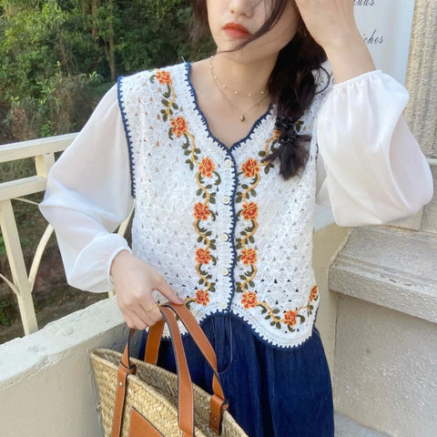 Woman wearing a white blouse with floral embroidery and dark pants, holding a straw bag outdoors.