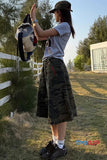 Person in camouflage shorts and cap holding a bag outdoors with a fence and trees in the background.