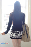 Person wearing a navy sweater and patterned skirt with a beige bag, standing by a window.