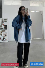Person wearing a denim jacket, white shirt, and black pants in a minimalistic room.