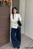 Woman in a white coat and blue pants standing on a wooden deck with a neutral wall background.