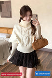 Woman taking a mirror selfie wearing a white cable knit sweater and black polka dot skirt, holding a brown handbag.