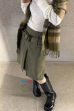 Person wearing a plaid scarf, white shirt, green skirt, and black boots against a concrete wall.