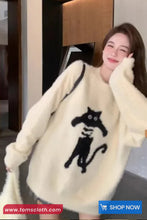 Woman wearing a white sweater with a black cat design, standing indoors.