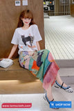 Woman sitting on a bench wearing a white t-shirt with a cat graphic and a colorful skirt, with 'www.tomscloth.com' displayed.