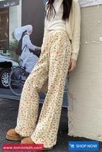 Person wearing a cream sweater and floral pants leaning against a wall with a blurred background.