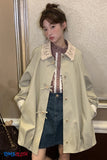 Person wearing a light green trench coat with a plaid shirt and denim skirt, standing against a neutral wall.