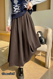 Person wearing a dark blue cardigan with white patterns, gray shirt, brown long skirt, and black shoes in a room.