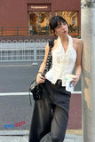 Woman in a white sleeveless top and black pants leaning against a wall on a street.