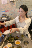 Woman preparing food at a counter with various dishes and a colorful wall in the background