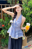 Woman in a blue top and dark pants holding a drink, standing outdoors with greenery.