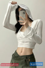 Person wearing a white long-sleeve top and green pants, holding a phone with a plain background.