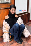 Woman sitting on a bench wearing a black off-shoulder sweater and blue jeans, with a white fur coat draped over her shoulders. 'TOMS CLOTH' logo is visible.