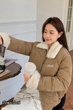 Woman in a brown jacket with white fur trim holding a cup indoors.
