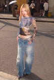 Woman in a patterned top and jeans standing on a street with people in the background.