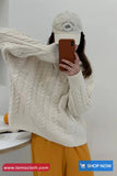 Person wearing a cream cable knit sweater and white cap, taking a selfie.
