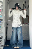 Person wearing a white floral blouse and blue jeans in a room with shelves and a bed.