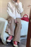 Person wearing a white cable knit sweater, beige pants, and red high-top sneakers sitting on a bed.