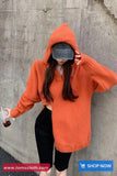 Person wearing an orange hoodie and black shorts against a gray concrete wall, holding a drink.