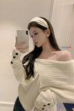 Woman wearing an off-shoulder white sweater with black buttons, holding a phone.