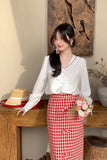 Woman in a white blouse and red checkered skirt standing in a room with wooden furniture and decorative items.
