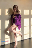 Woman in a purple off-shoulder dress standing against a wall with sunlight casting shadows.