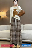 Person wearing a white sweater with a teddy bear design and plaid skirt, holding a brown handbag in a living room.