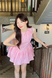 Woman in a pink checkered dress standing indoors with a staircase in the background